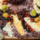 A board on a table filled with many, many different kinds of sliced meats, cheese, fruits, vegetables, and spreads.